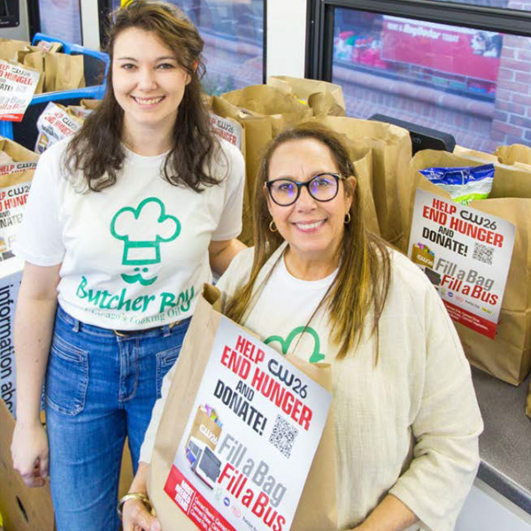 Two woman with butcher boy branded shirts on surrounded by bags that read, "Hep end hunger and donate. Fill a bag, fill a bus."