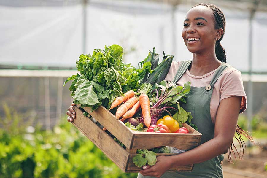 Woman carrying a wooden box filled with an array of different vegetables.